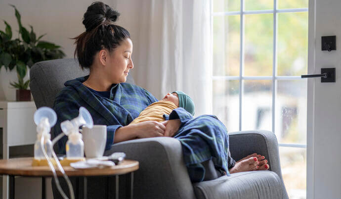 A woman holding her baby and her breast pump is sitting on the table beside her.